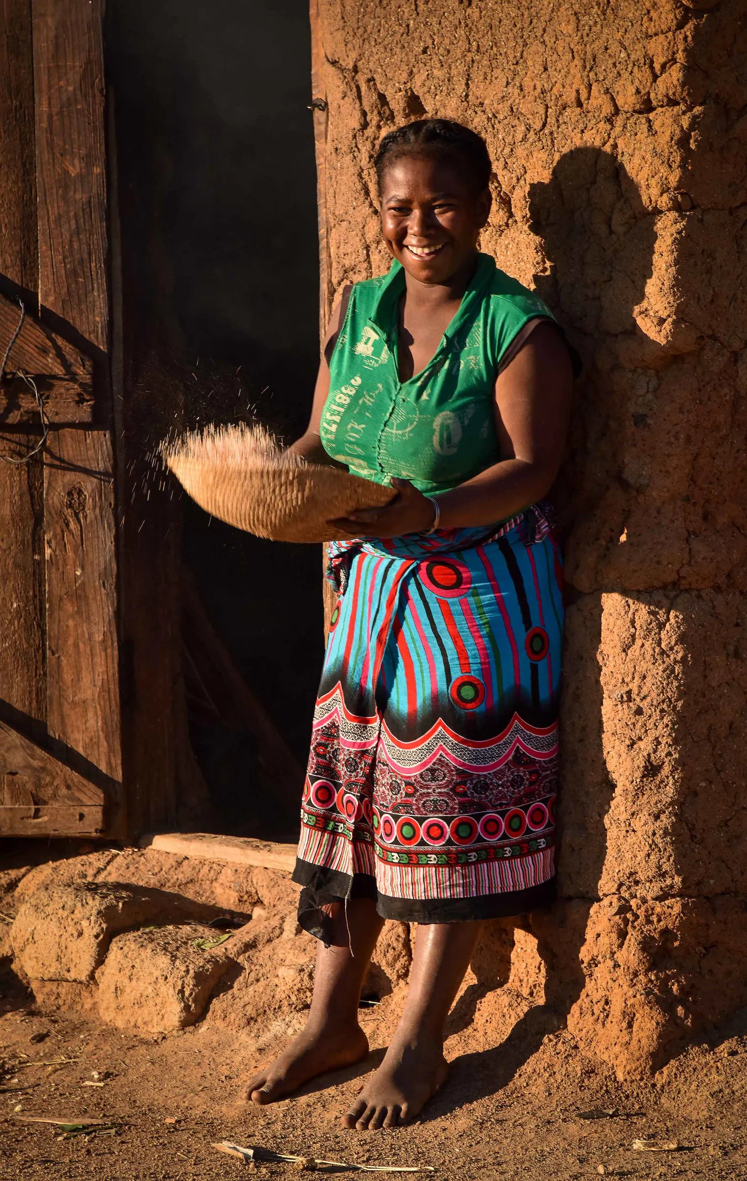 A smiling woman winnows rice with a sahafa basket in her doorway